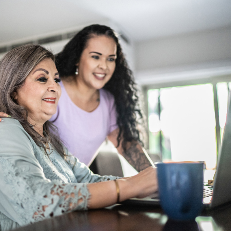 A Hispanic senior woman reviews information on a laptop computer while her daughter watches over her shoulder.