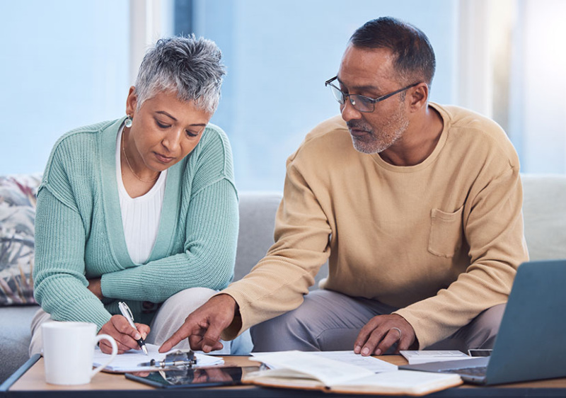 A senior man and woman of color are seated at a couch and examining documents set out on a coffee table. The woman is signing a document.