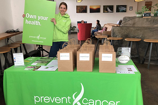 A young woman wearing a bright green sweatshirt holds up a green sign that reads, “Own your health.” She is standing behind a table that has a bright green table cover that features the Prevent Cancer Foundation logo on the front in white. The table has materials, gift bags and a bowl for donations.