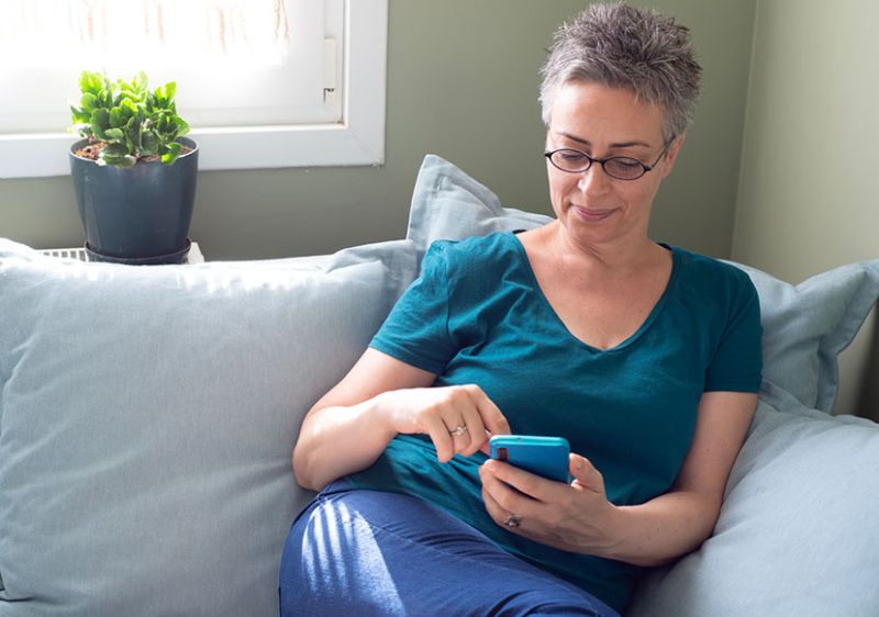 A woman in her 40s is curled up on a cozy couch with a phone in her hand. She is next to a window, which is shining bright into the room. The woman has short grayish hair, is wearing glasses and appears to be scrolling content on the phone.