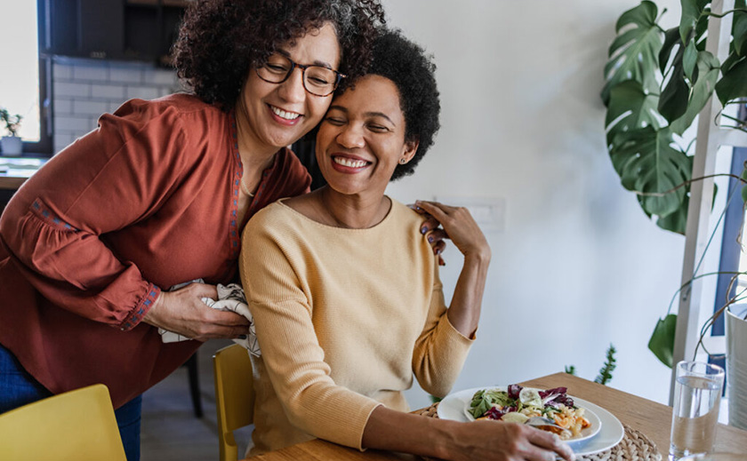 A close-up image of two women in their 40s or 50s in a dining room. One woman is seated at the table. The other woman is behind her and leaning down to embrace the other woman's shoulders. Both women are smiling and eyes are closed indicating contentment.