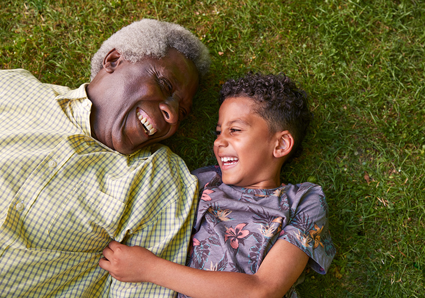 A senior Black man is laying on his back in the grass next to a young Black boy who appears to be his grandson. They are tilting their heads toward each other and smiling.