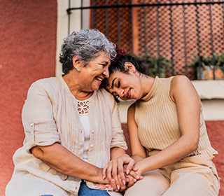 A young woman with brown skin and dark hair lays her head on the shoulder of an older woman who appears to be her grandma. Both are smiling and look content.