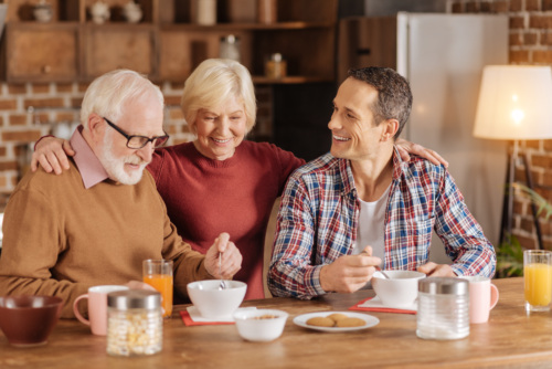 An elderly woman hugs her adult son and husband while they sit at the kitchen table eating oatmeal.
