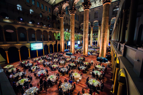 A room filled with round tables, decorations and mood lighting for a gala event.