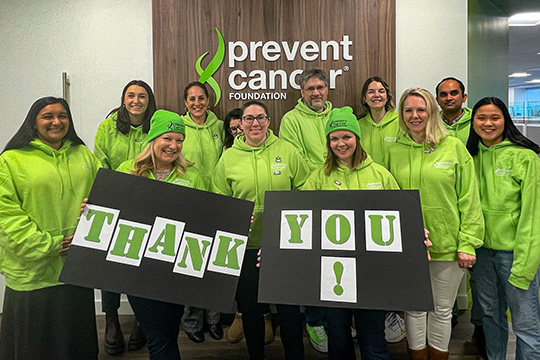 Ten adults gather behind a large sign that reads, “thank you.” There are two males and nine females, all wearing bright green sweatshirts. You can see a large Prevent Cancer Foundation logo mounted on the wall behind them.