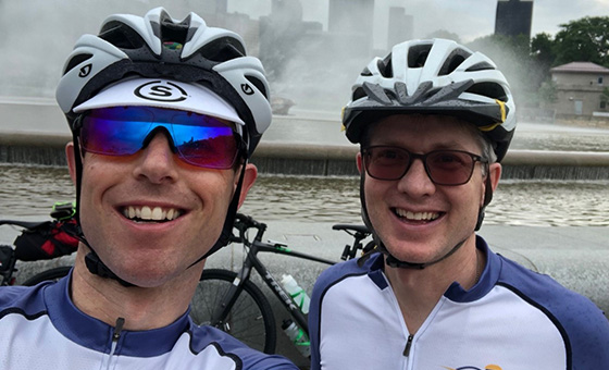 A close-up of two adult white males wearing biking gear and sunglasses in front of a fountain.