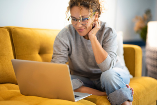 A woman wearing a grey sweater, glasses and blue jeans sits on a yellow couch, looking at her laptop screen.