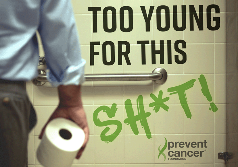 A man in his 40s wearing dress clothes is facing away from the camera in front of a toilet in a stall, holding a roll of toilet paper in his right hand. There is text painted on the wall tiles next to the toilet that reads, “Too Young for This Sh*t!”