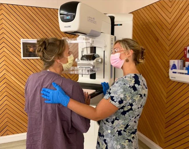 Two women wearing masks stand at a mammogram machine. One of them, a health care provider, helps the other, a patient dressed in a medical gown, get into position for her breast cancer screening.
