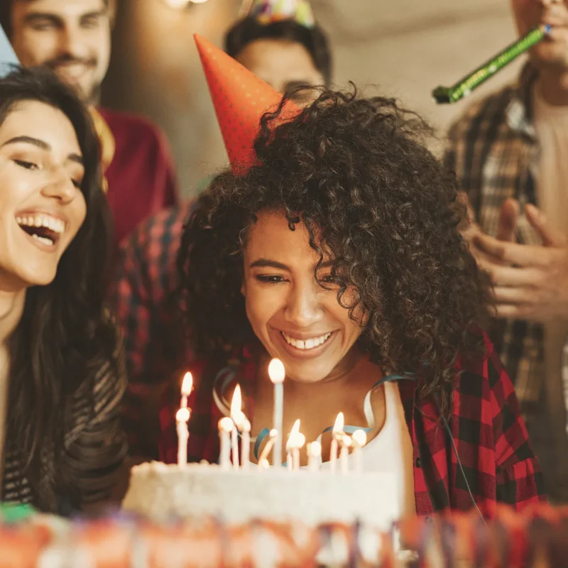 A young woman sits with a cake in front of her and lit birthday candles. She is smiling and wearing a party hat. There are smiling and clapping people surrounding her.